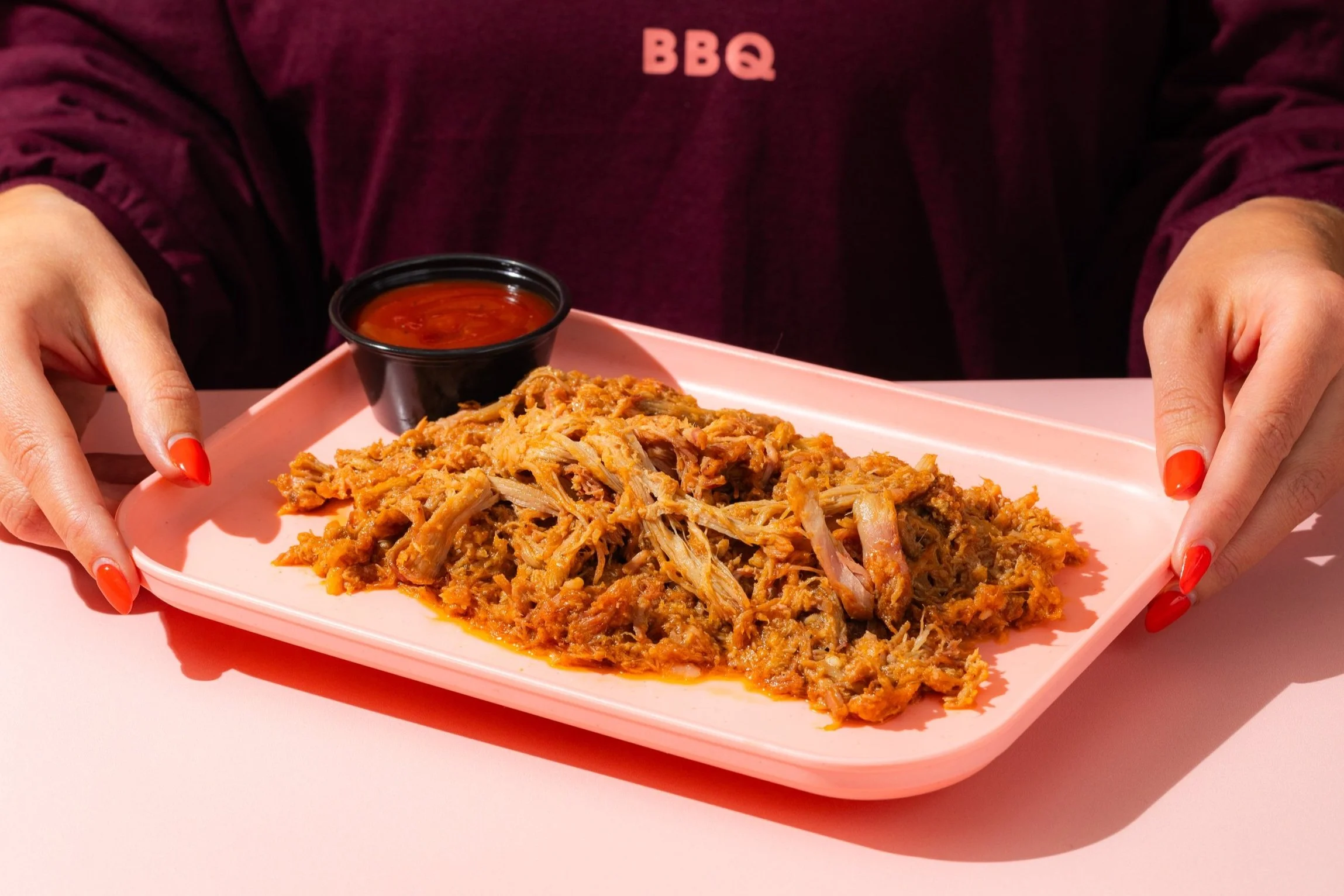 Hands holding a pink tray of Luv Que pulled pork with sauce